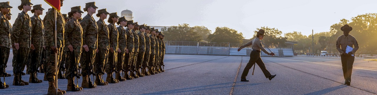A Marine Corps Drill Instructor marches on the parade deck in front of a platoon of Marine Corps Recruits. 