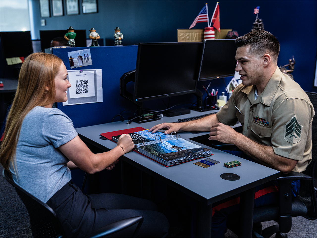 Marine recruiter in service uniform meeting with prospective recruit reviewing enlistment materials at desk.