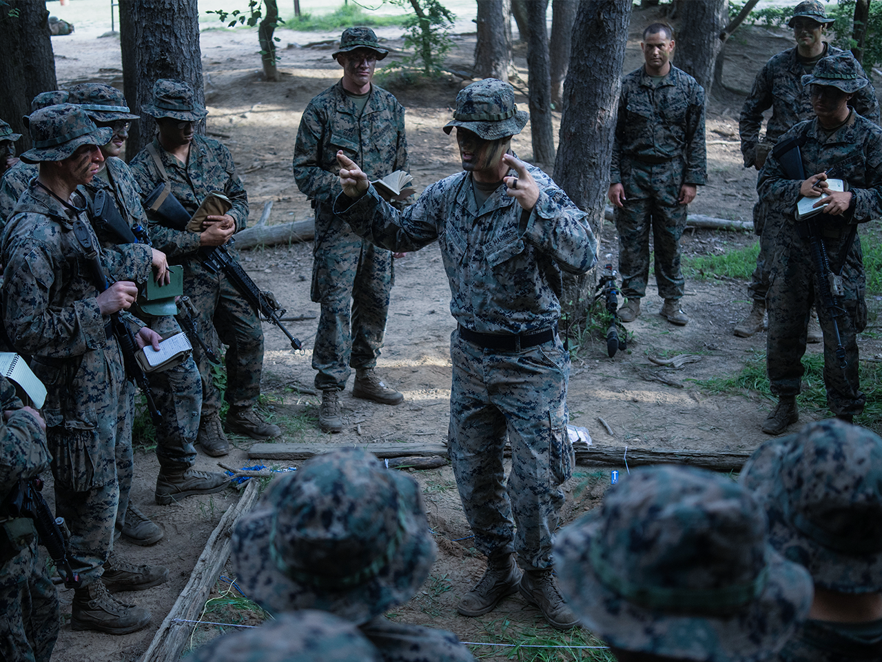 Marine instructor providing tactical briefing to group of Marines in camouflage during field training.