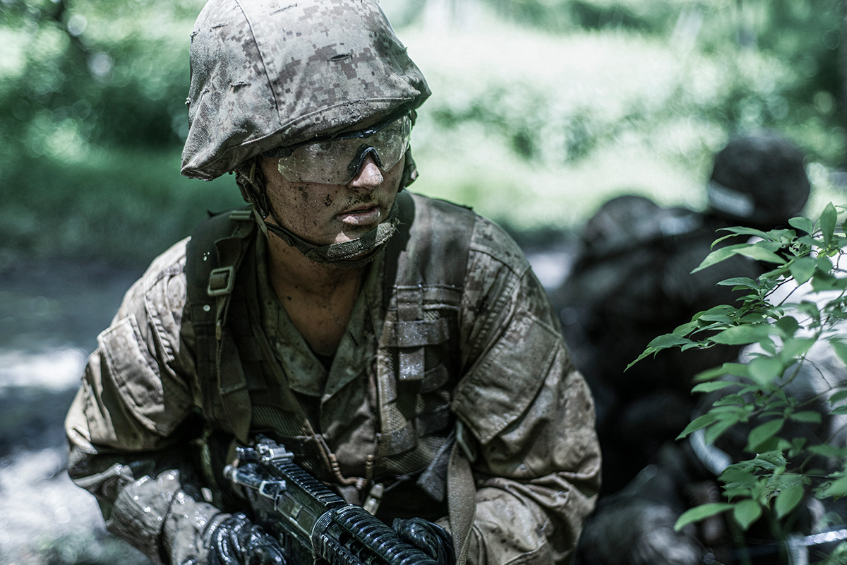 Officer candidate covered in mud holds rifle during combat endurance course training in wooded terrain.