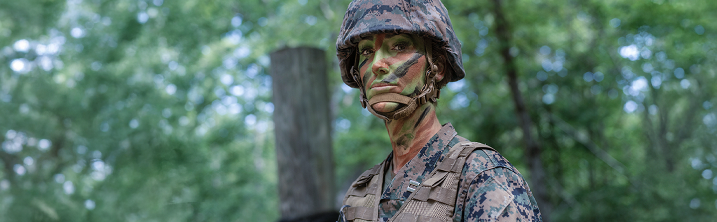 Marine officer with camouflage face paint and helmet in wooded training environment.