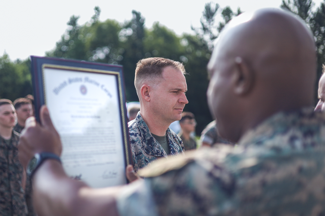 Marine receiving framed certificate or diploma during outdoor graduation ceremony with audience.