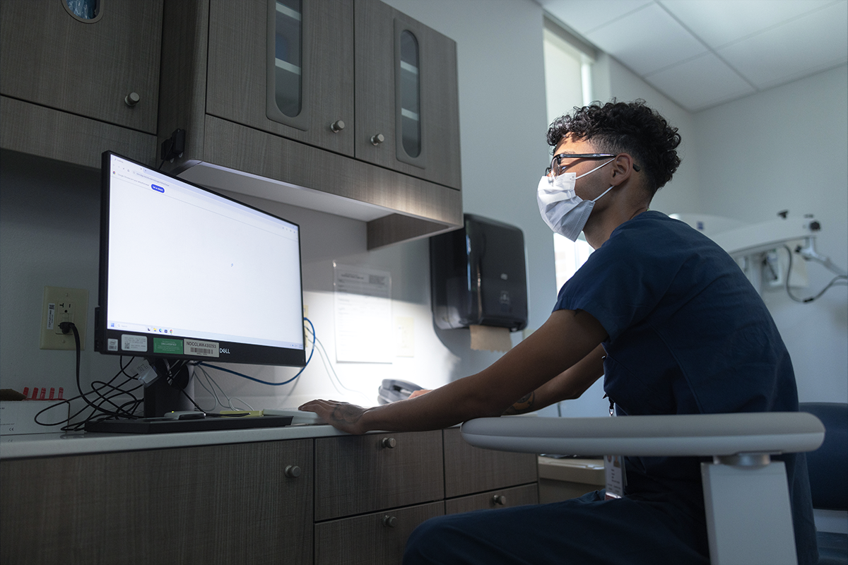 Healthcare provider in mask and scrubs using computer workstation in modern medical facility examination room.