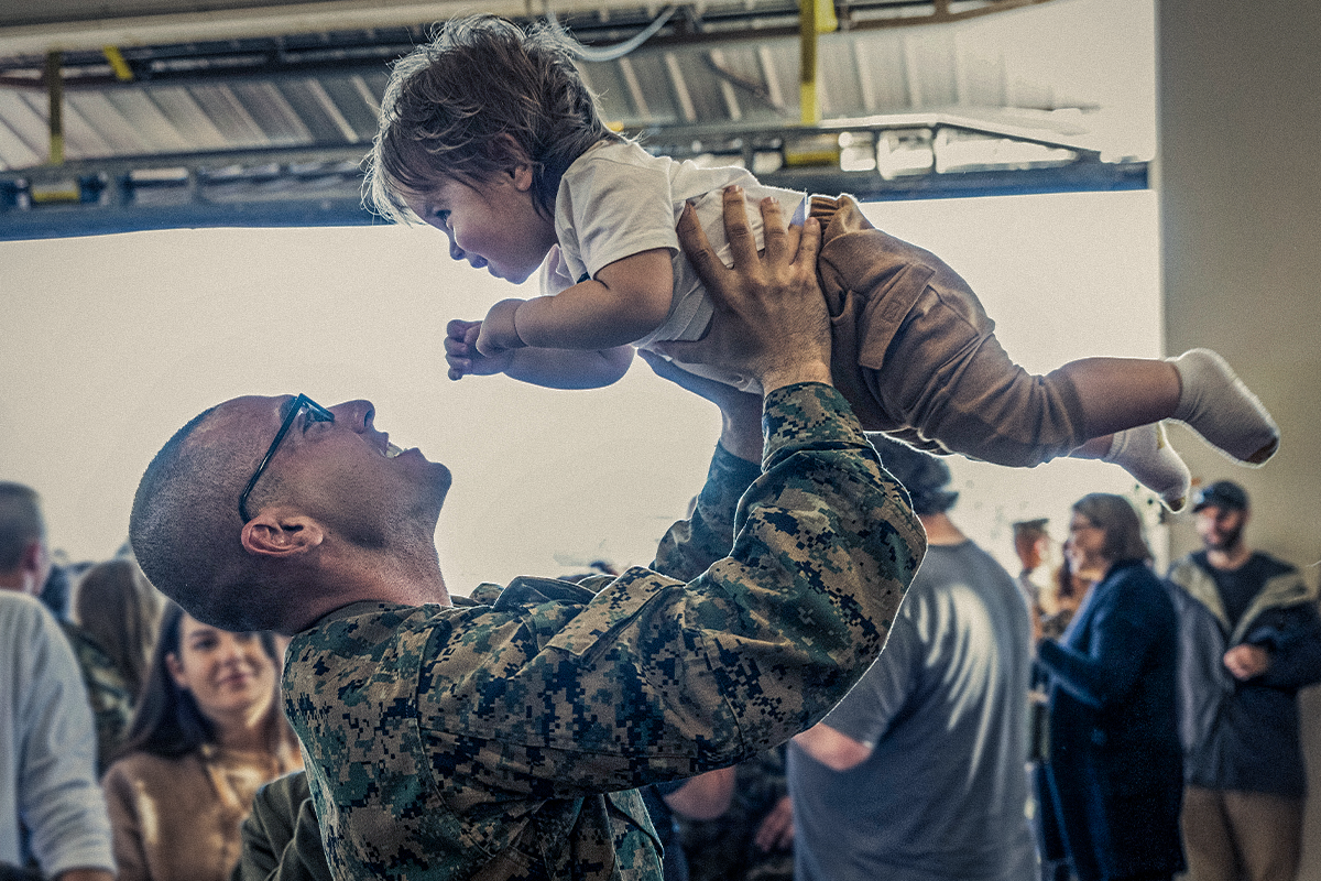 Marine in woodland camouflage lifting young child in hangar during family reunion with other Marine families present.