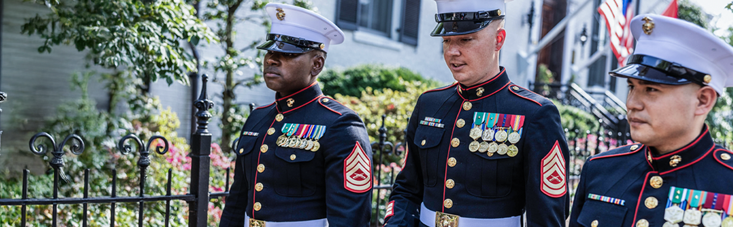 Marine officers in dress blue uniforms with ribbons and white covers outside building.
