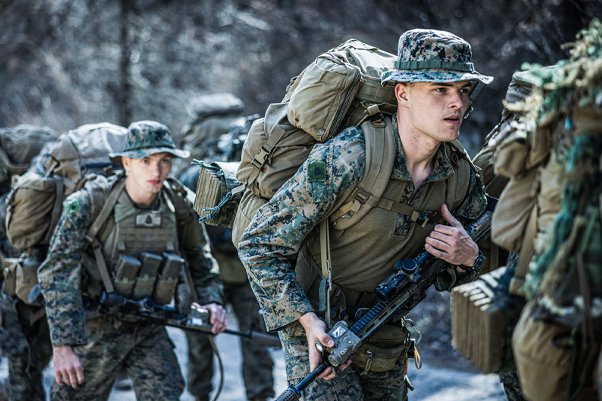 Marines in camouflage uniforms carrying heavy rucksacks and rifles during cold weather training exercise.
