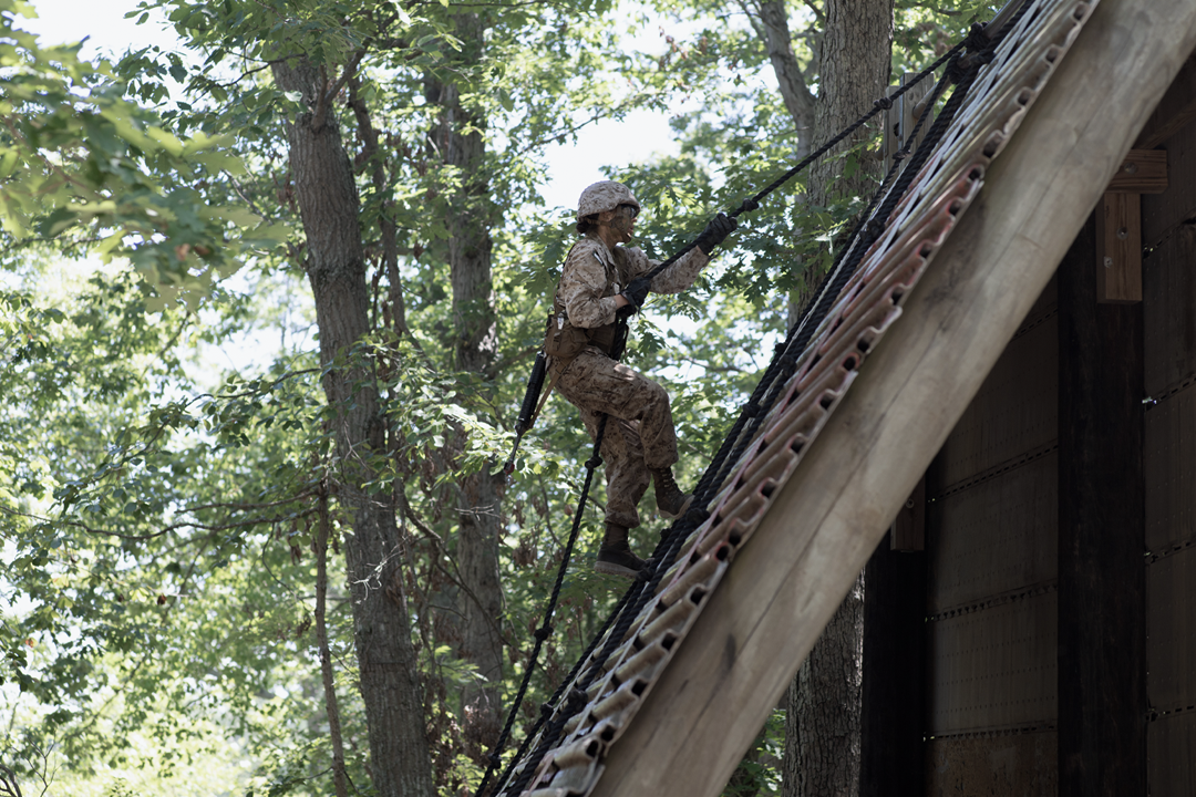 A Marine uses a rope to climb up a steeply angled wall. They are completing a combat field course.