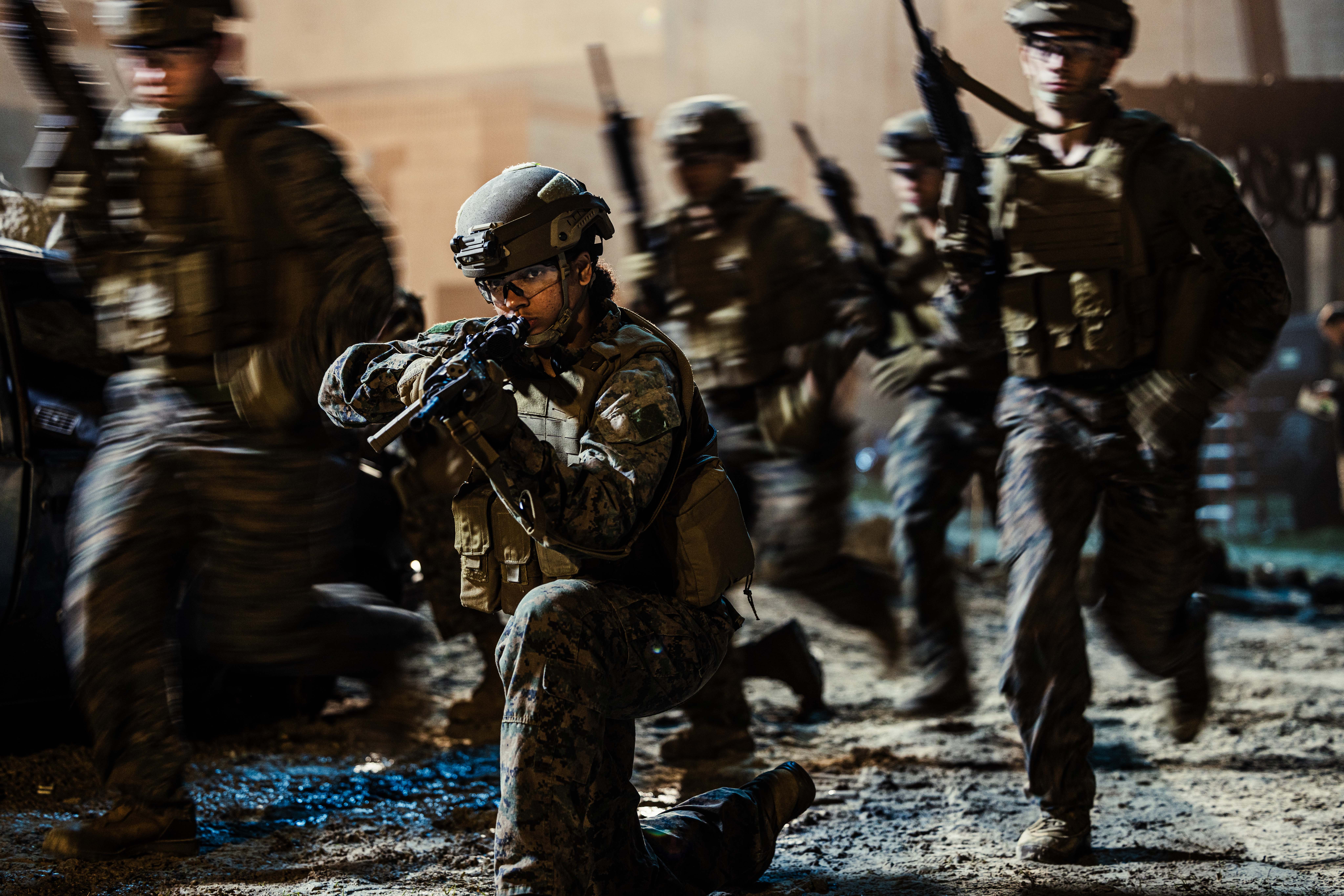 A Marine takes a knee and aims a rifle while fellow Marines in full combat gear advance around them during a low-light operation.