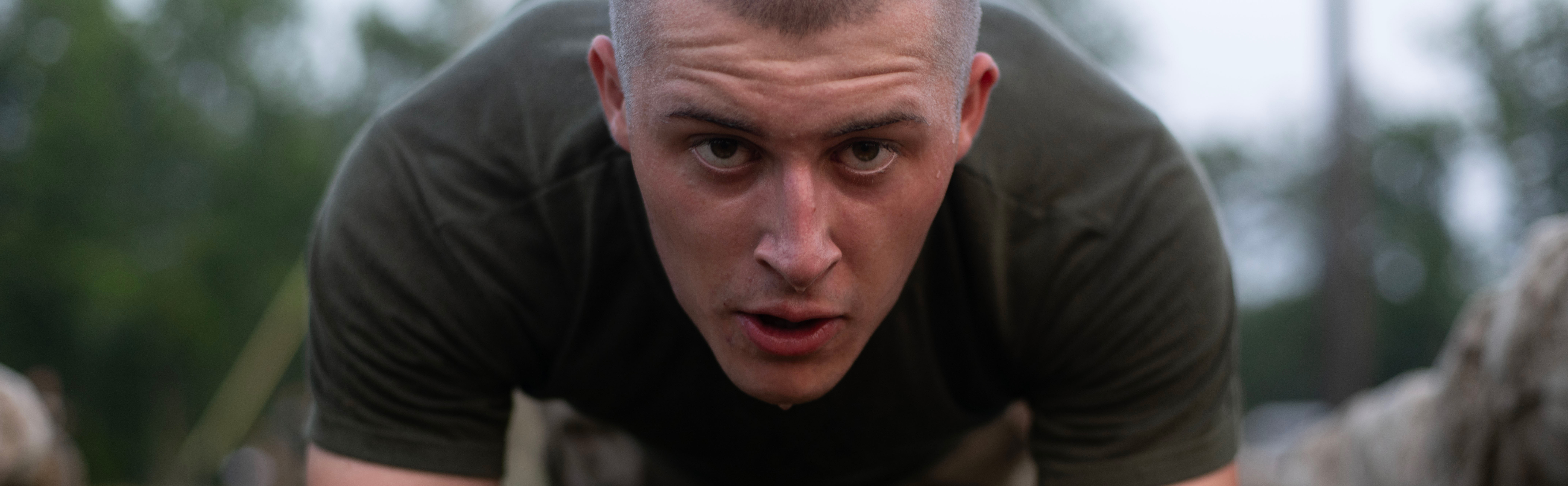 A Marine recruit with a shaved head completes a plank. He is looking straight into camera. 