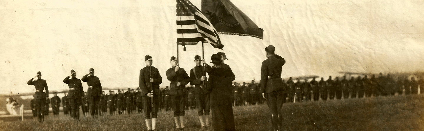 Historic black and white photo of early Marines standing at attention with American flag and battle colors during ceremony.