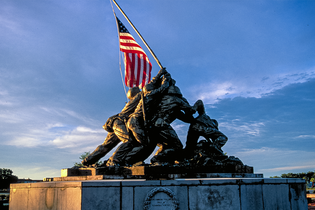 Iwo Jima Memorial statue showing Marines raising American flag, illuminated against dramatic evening sky.
