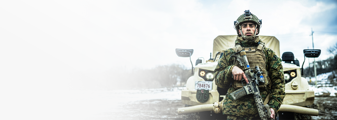 Marine in combat gear with rifle standing ready beside armored vehicle during field operations.