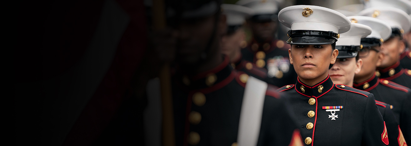 Marine in dress blue uniform with white cover and red trim standing at attention during formal ceremony.