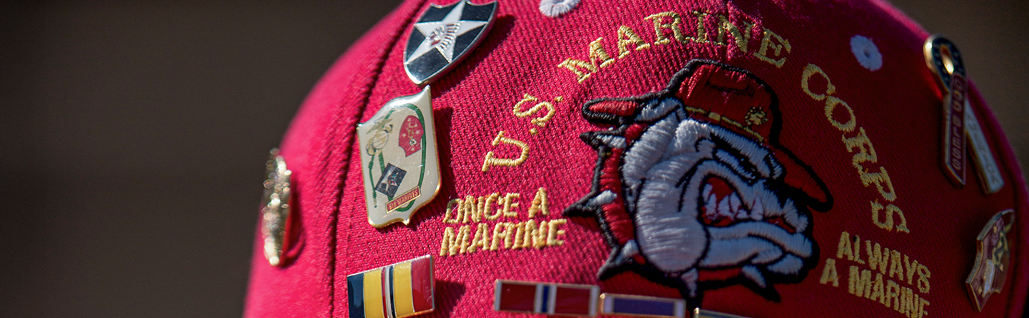 A red USMC hat covered in pins and ribbons with a bulldog mascot on the front and embroidery spelling "Once a Marine Always a Marine." 