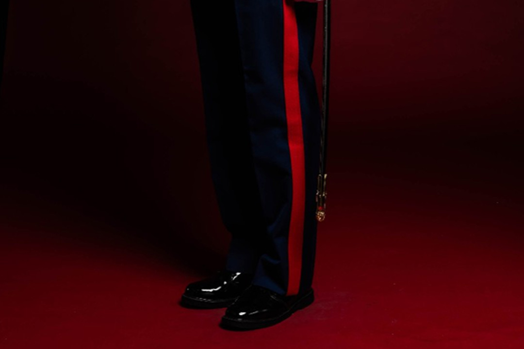 A Marine in dark blue uniform with red stripe, shiny black shoes, and a hanging sword against a dark red background.