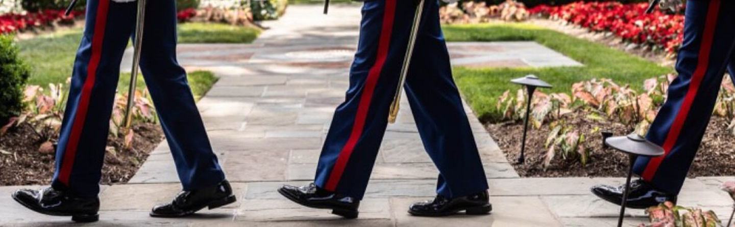 Three Marines with red stripes and shiny black shoes walk on a paved path next to gardens with green grass and red flowers.