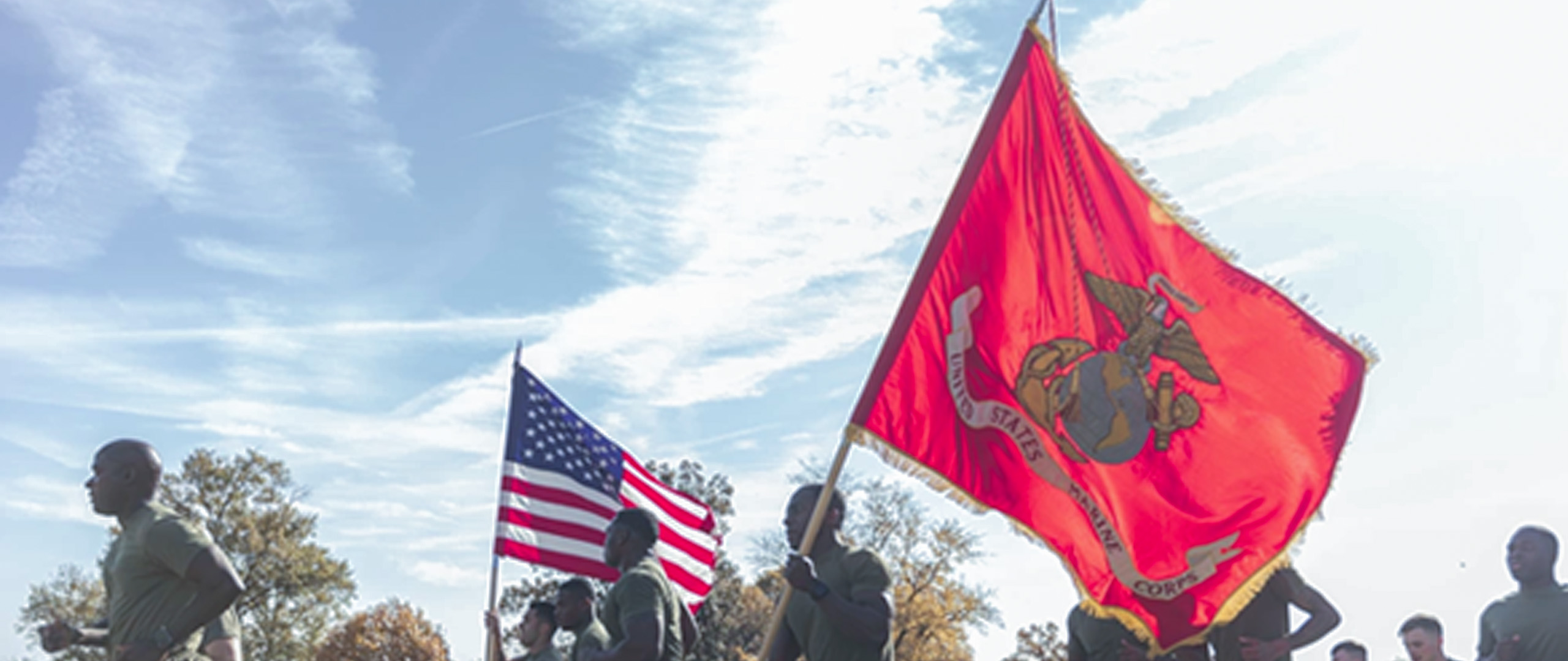 A group of Marines run across a grassy field, carrying the US flag and a large red Marine Corps flag featuring the eagle, globe, and anchor.