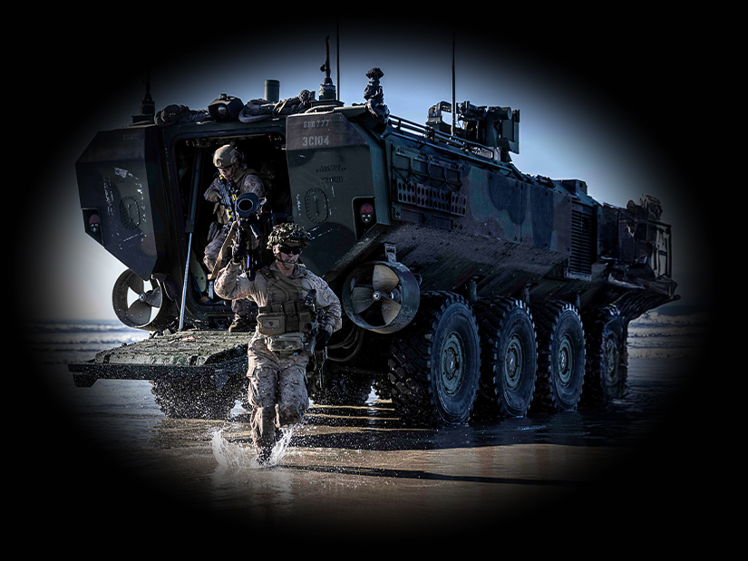 Two Marines run out the back of an amphibious assault vehicle. They run across wet sand on the beach. 