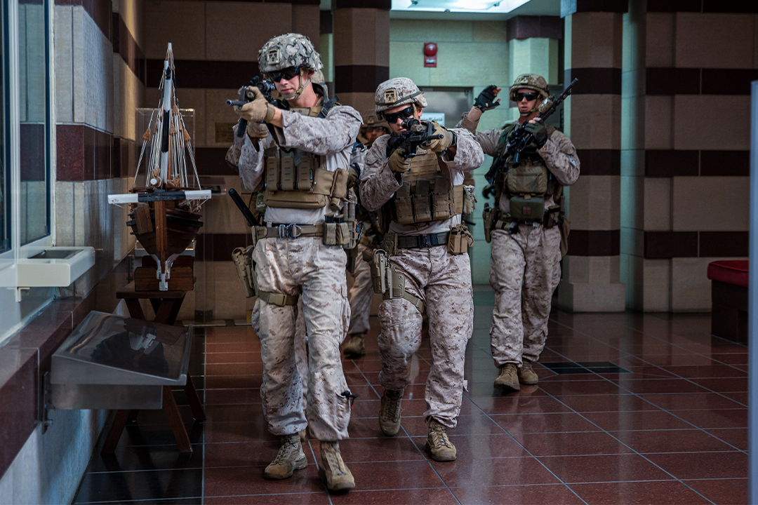 Marines in camo and with rifles drawn walk through the lobby of a government building. 