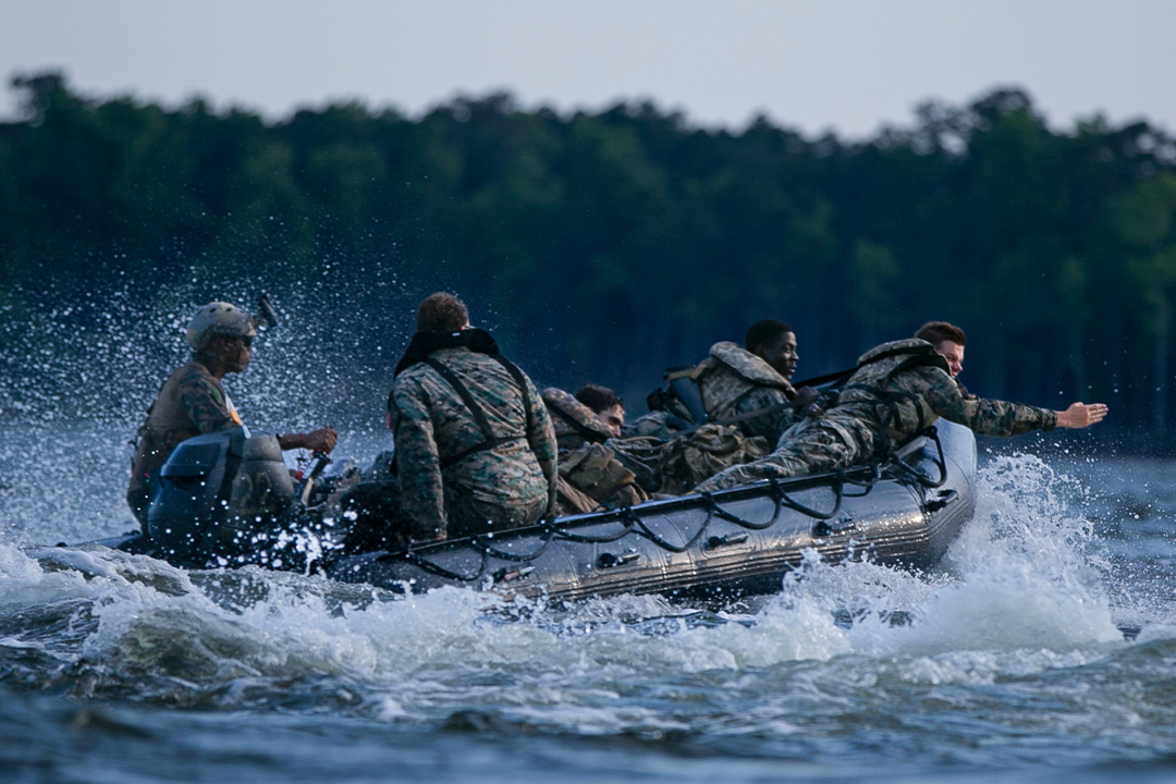 5 reconnaissance Marines ride aboard a small boat. Surf splashes up around them as the boat moves through the water. 