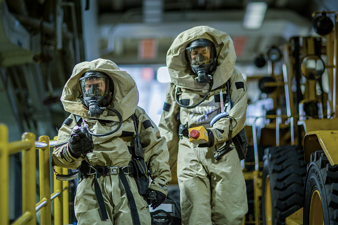 Two chemical, biological, radiological, and nuclear Marines walk among vehicles with full protective suits and detection equipment.