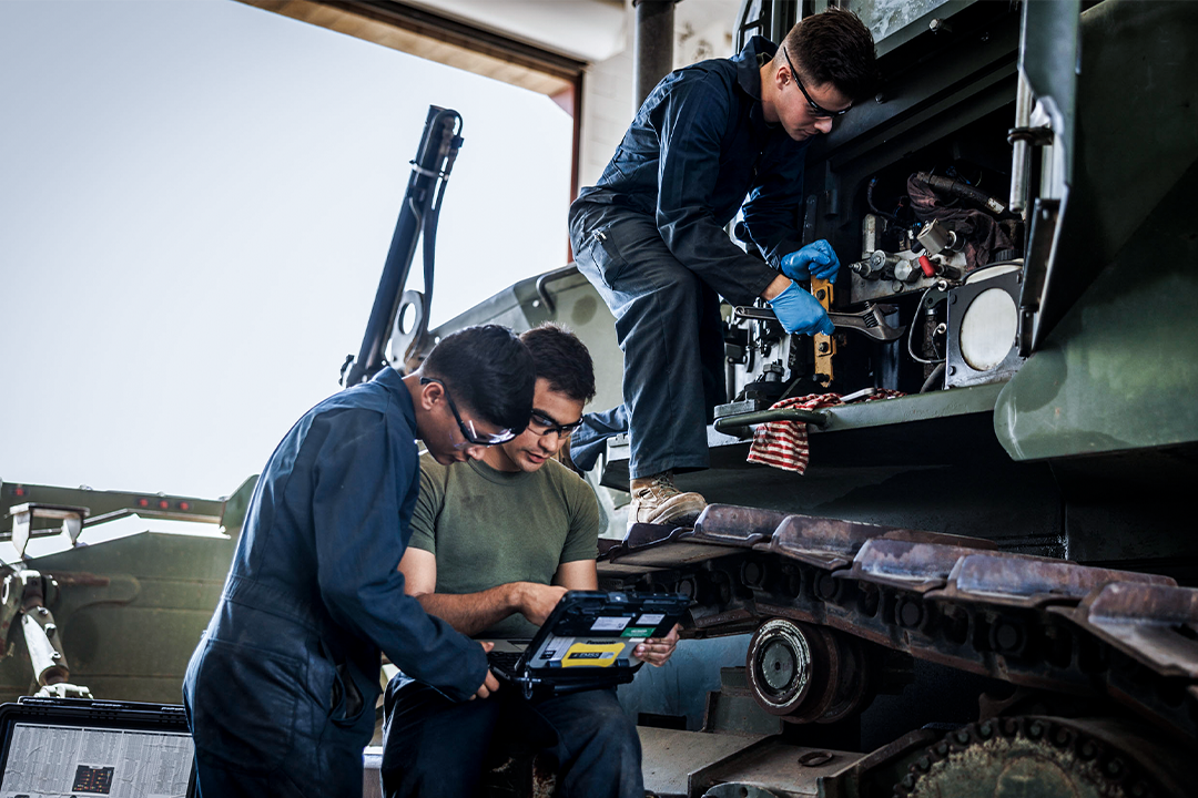 Three vehicle mechanic marines work on a large USMC vehicle in a garage. 