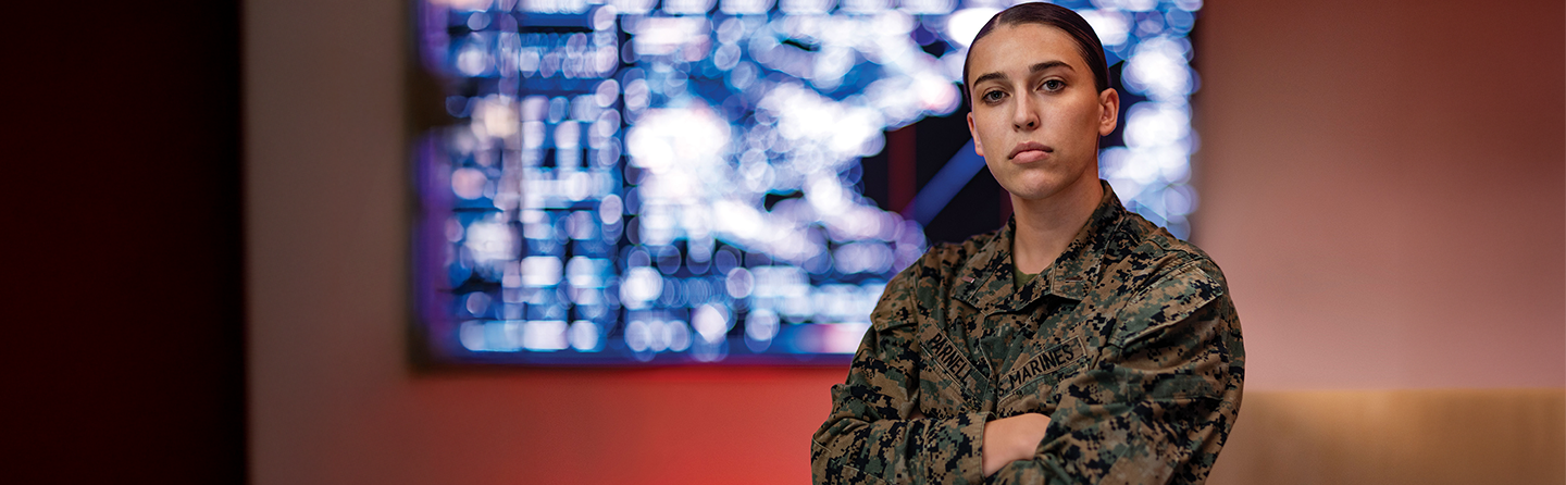 First Lieutenant Annie Parnell stands with arms crossed in utility uniform with digital displays in background.