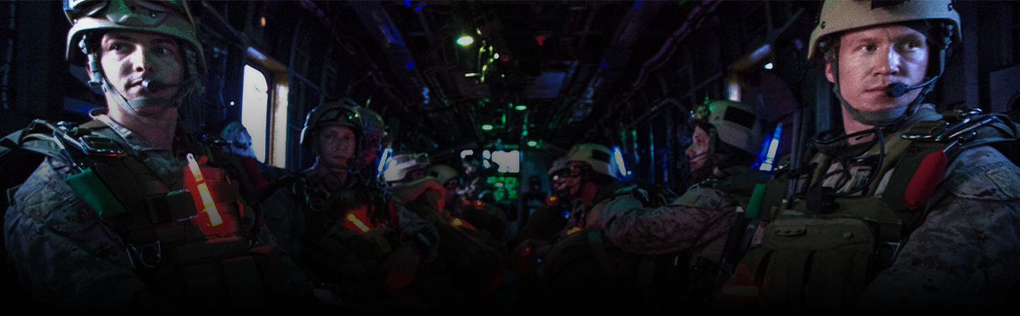 Several Marines in combat military gear sit inside a dark transport helicopter, illuminated by red and green lights.