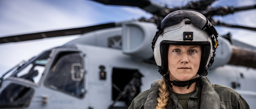 Captain Kricket Harper in flight helmet standing in front of military helicopter on flight line.