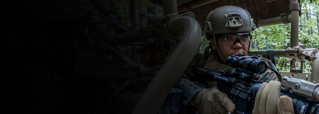 A Marine riding in a MRZR tactical vehicle brings their rifle up to their face while training in a forest clime.