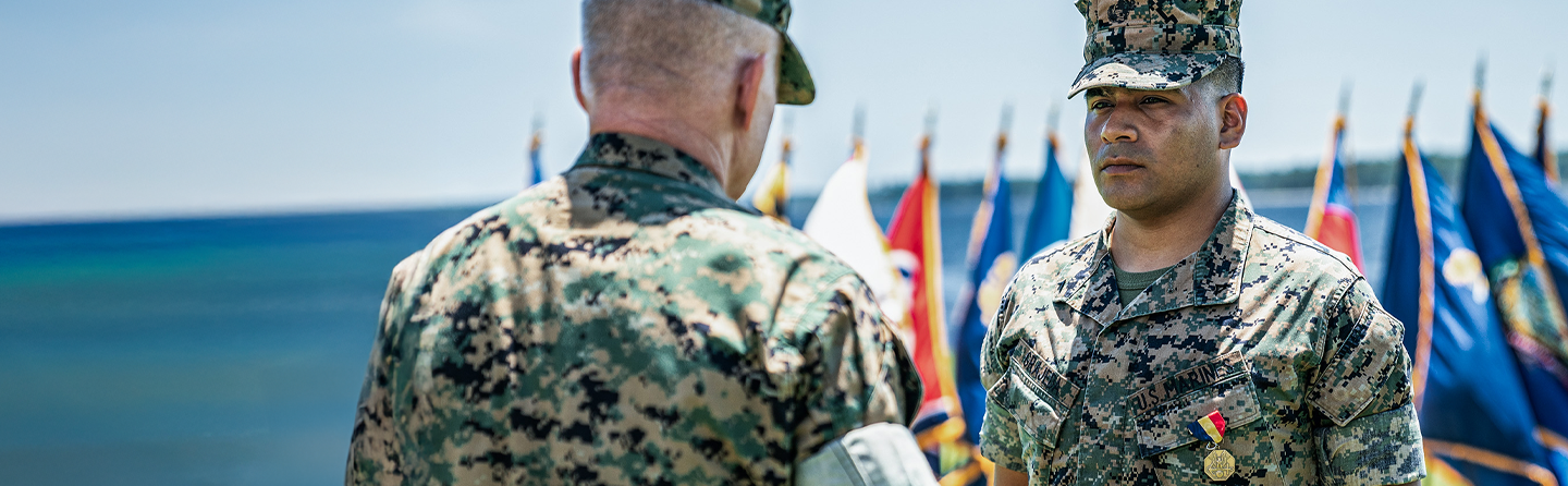 Lance Corporal Kevin Grajeda stands at attention in service uniform with medal during outdoor ceremony with flags.