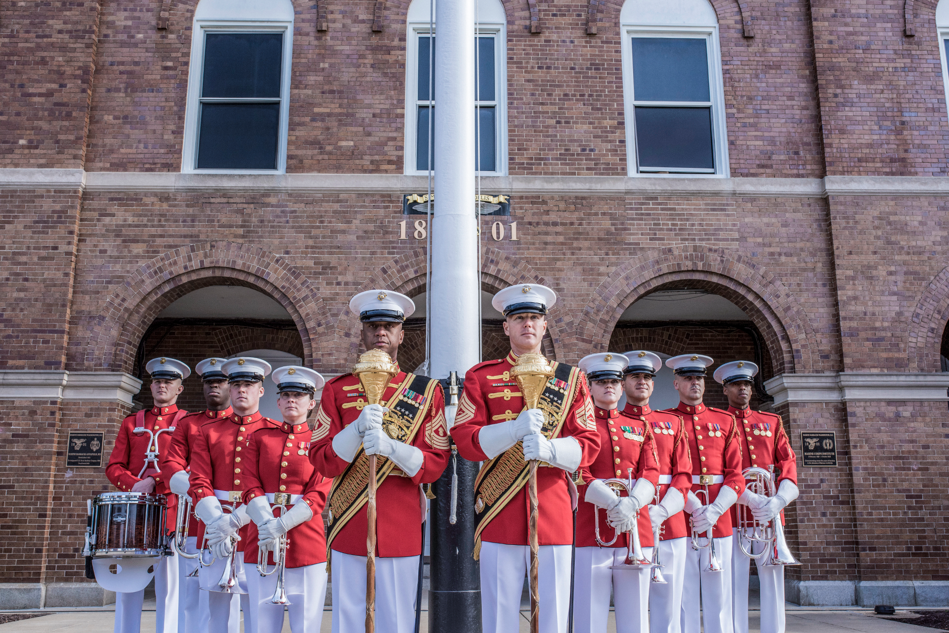 Marine band members in red dress uniforms stand in formation holding instruments in front of brick building.