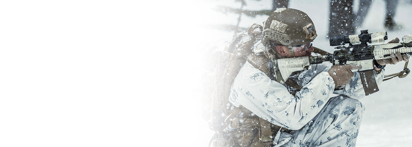 Marine in snow camouflage with scoped rifle conducting arctic warfare training during winter conditions with blowing snow.