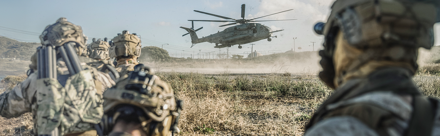 Military helicopter landing with dust cloud while Marines in combat gear observe special operations training exercise.